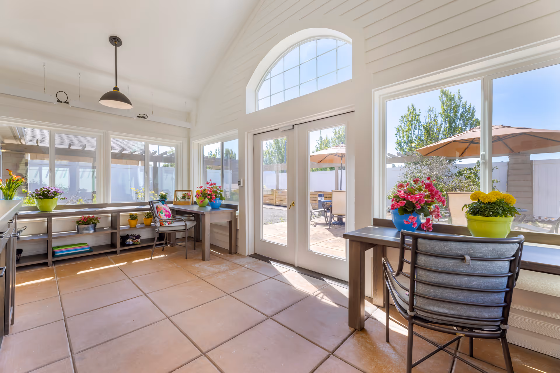 Bright sunroom with large windows and glass doors leading to an outdoor patio. The room has tiled floors, two desks with chairs, and colorful potted flowers on the desks and shelves. Outside, there are patio umbrellas and seating areas surrounded by greenery.