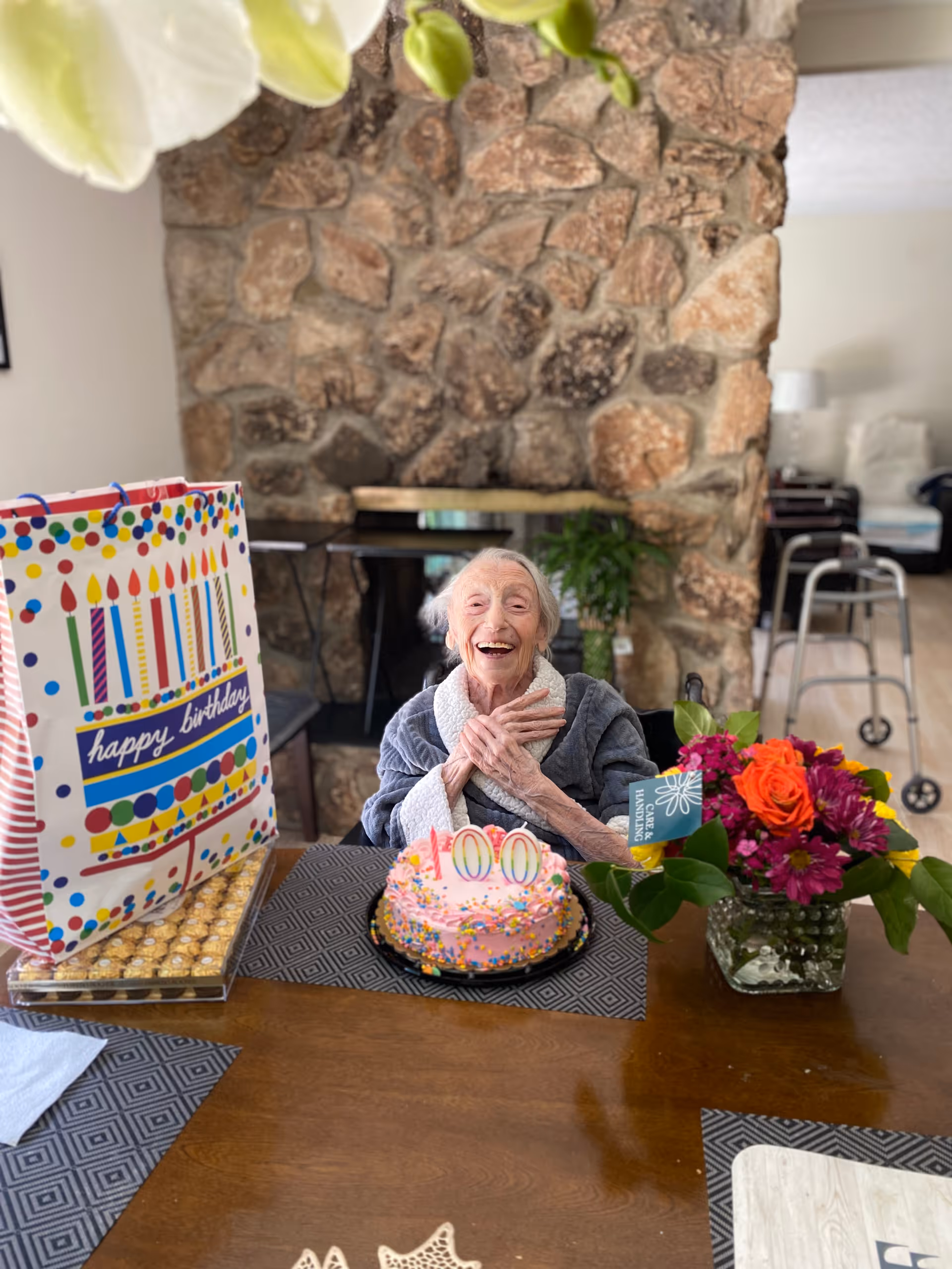 An elderly woman sitting at a wooden table with a pink birthday cake decorated with sprinkles and candles showing the number 100. She is smiling and has her hands crossed over her chest. On the table, there is a colorful birthday gift bag, a box of chocolates, and a vase with a bouquet of flowers. Behind her is a stone fireplace and a walker is visible in the background.
