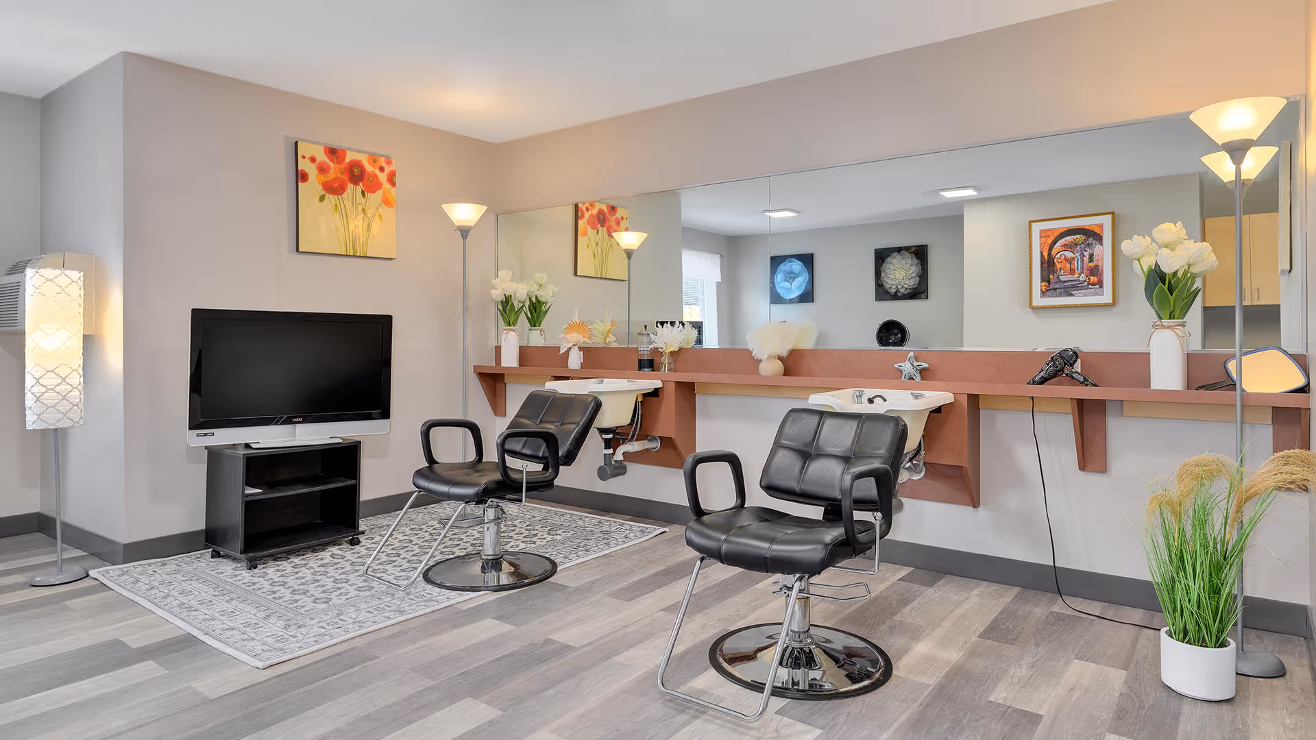 Interior view of a salon area in a senior living facility with two black salon chairs in front of a long mirror and counter with hair washing sinks. The room has light gray walls, a TV on a stand, decorative flowers, framed artwork, and modern floor lamps.