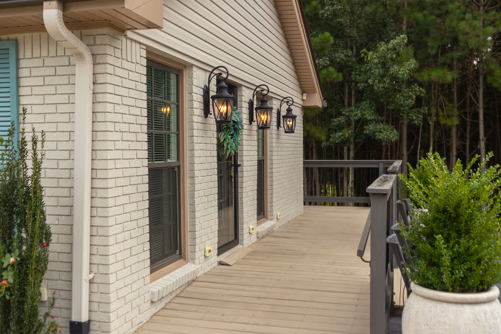 Wooden deck beside a light-brick house with three wall lanterns, windows, railing, and potted plants overlooking trees.