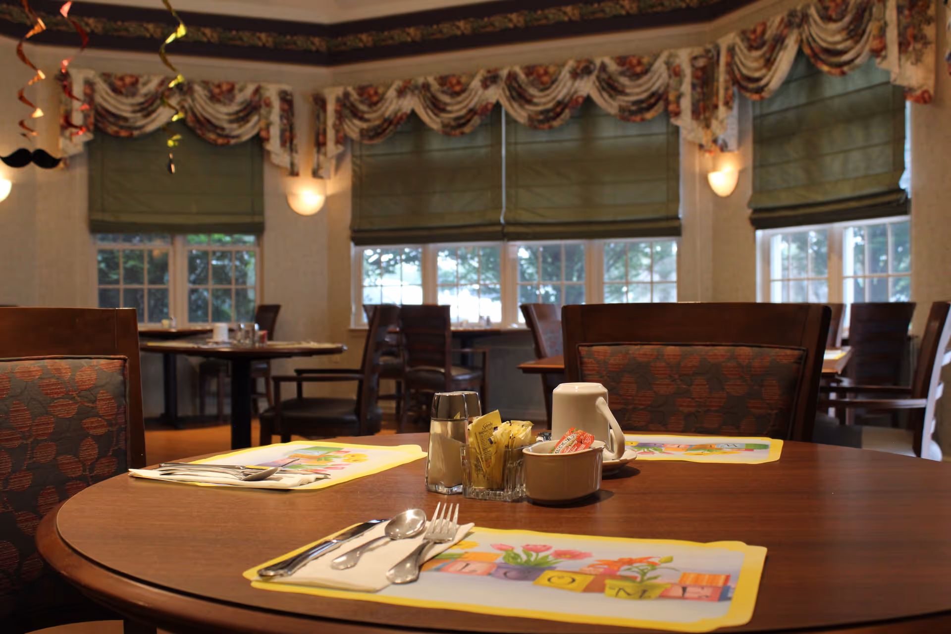 A dining room in Burton's Ridge Assisted Living with wooden tables and chairs. The tables are set with placemats, utensils, and condiments. The room has large windows with green Roman shades and floral valances, and soft wall lighting.