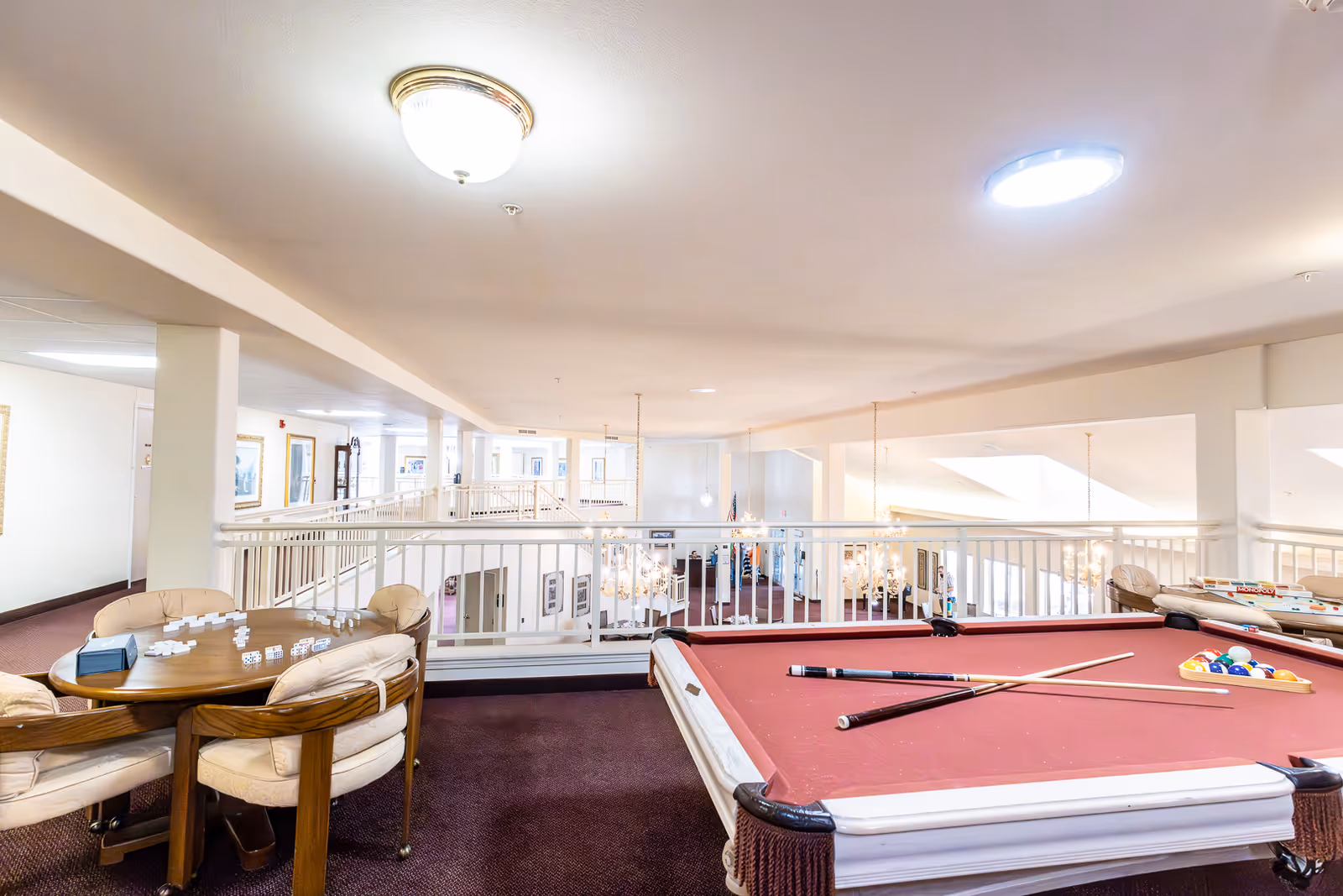 Interior view of a senior living facility's recreational area featuring a red pool table with cues and balls arranged, a round table with chairs and dominoes set up, white railings overlooking a lower level, and bright ceiling lights illuminating the space.