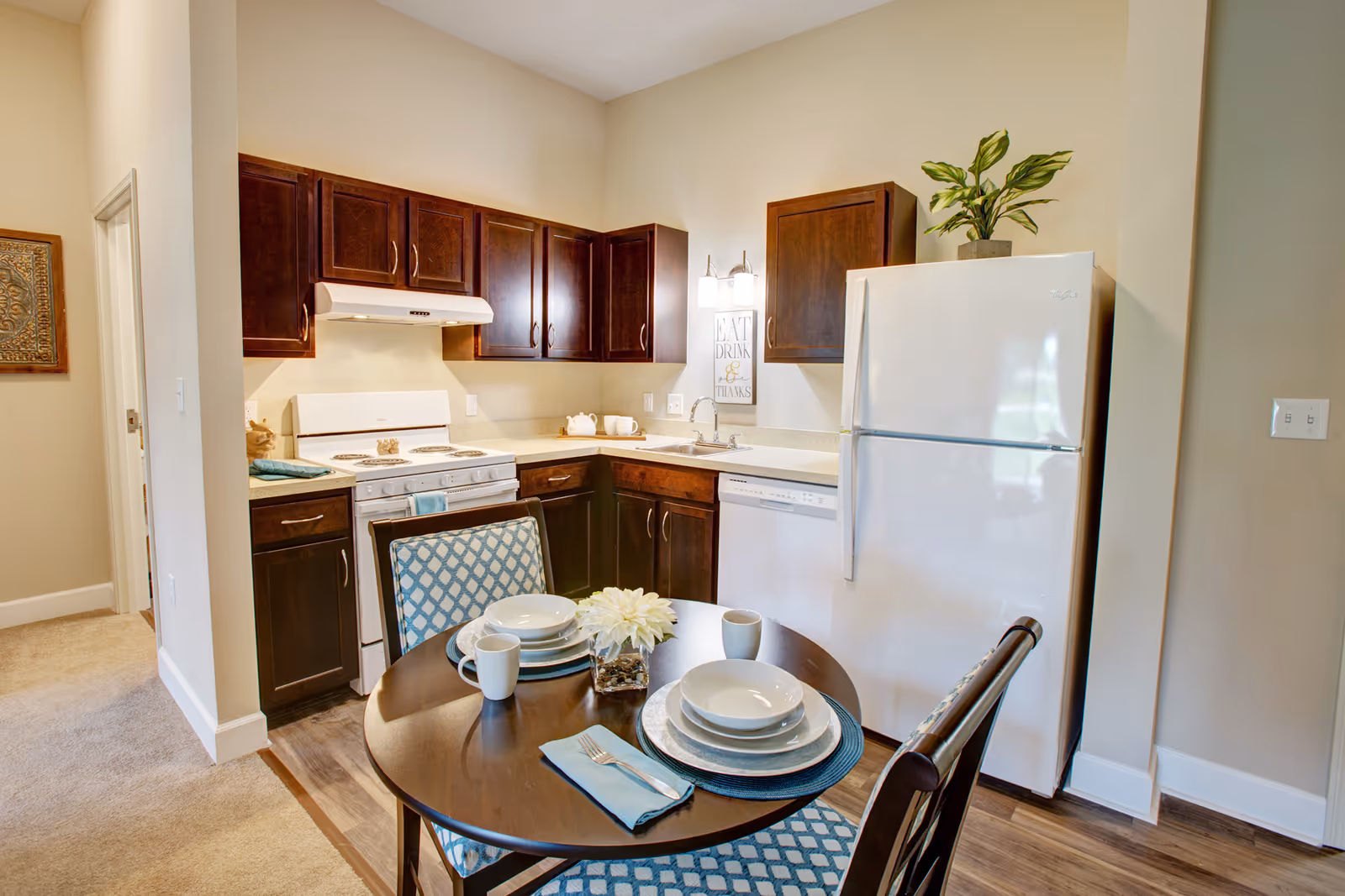 A compact kitchen area with dark wood cabinets, a white stove, white refrigerator, and a dishwasher. There is a small round dining table with two chairs, set with plates, bowls, mugs, and a small flower arrangement. A decorative sign on the wall reads 'EAT DRINK & GIVE THANKS'.