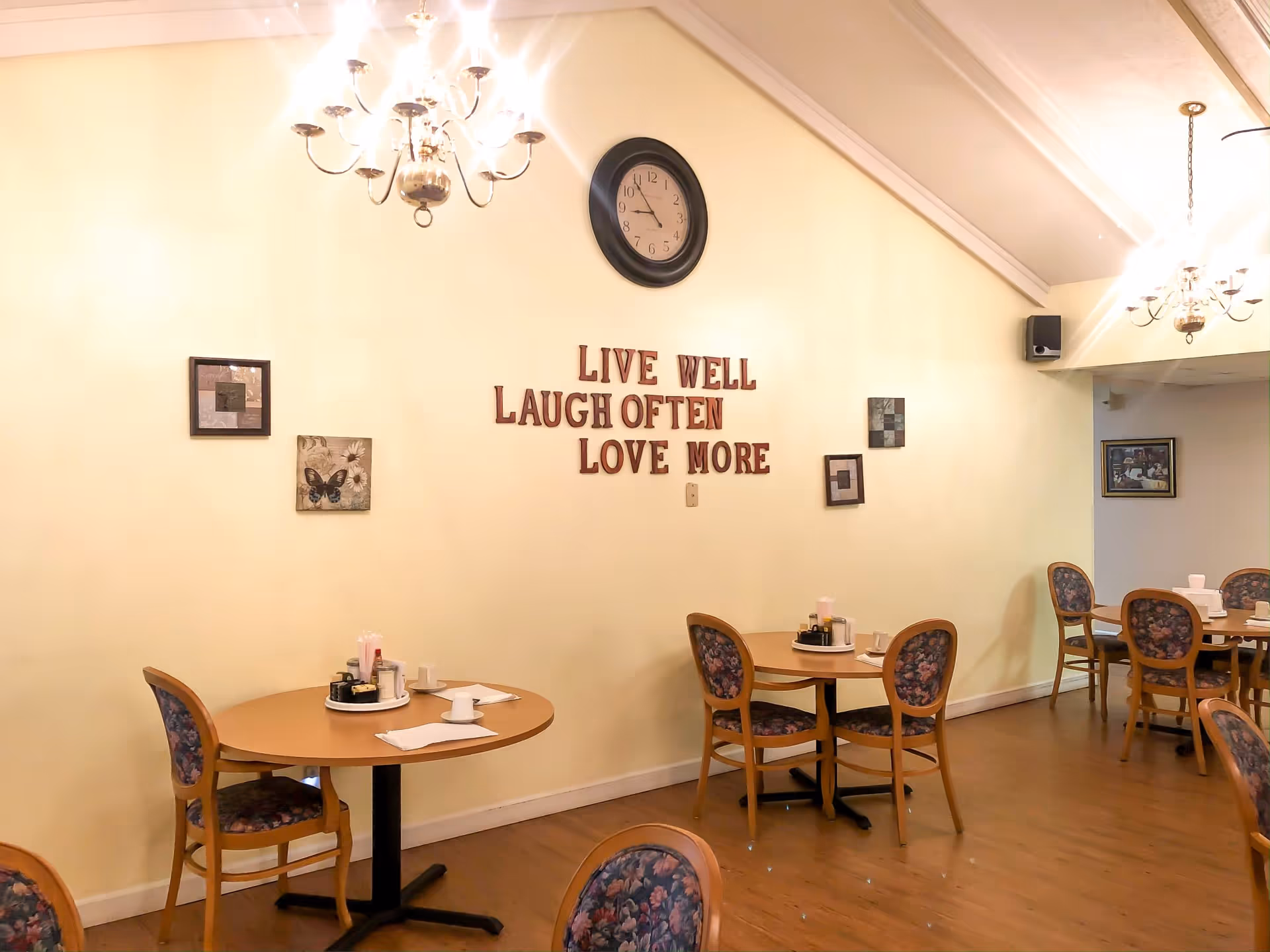 Interior view of a senior living community dining area with round tables and floral upholstered chairs. The wall features a clock and decorative text that reads 'LIVE WELL LAUGH OFTEN LOVE MORE' along with framed artwork. Two chandeliers hang from the ceiling providing warm lighting.