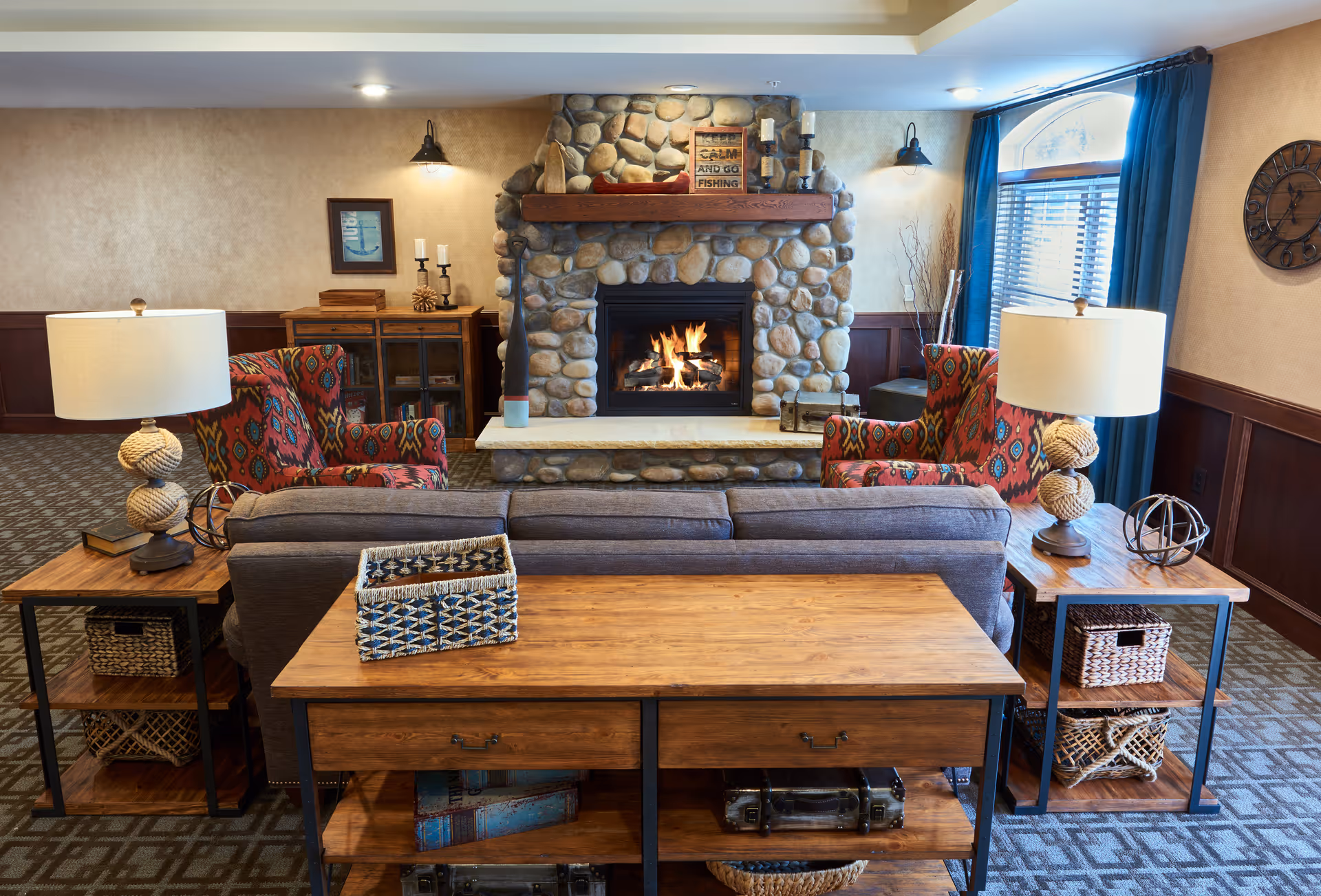 Cozy living room area with a stone fireplace in the center, two patterned armchairs on either side, a gray sofa facing the fireplace, wooden side tables with lamps, and a large window with blue curtains letting in natural light.