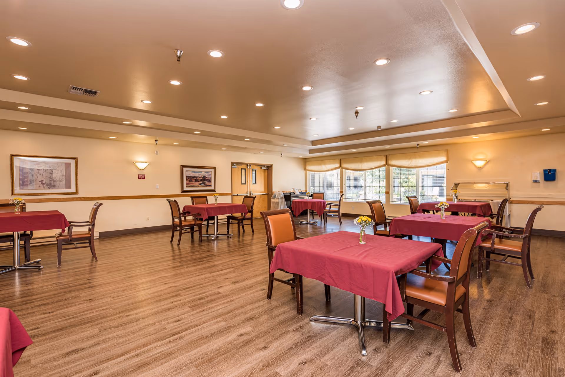 A spacious dining room with several tables covered in red tablecloths, each with a small vase of yellow flowers. The room has wooden flooring, beige walls with framed artwork, large windows with beige valances allowing natural light, and recessed ceiling lights. There are wooden chairs with brown cushions around the tables.