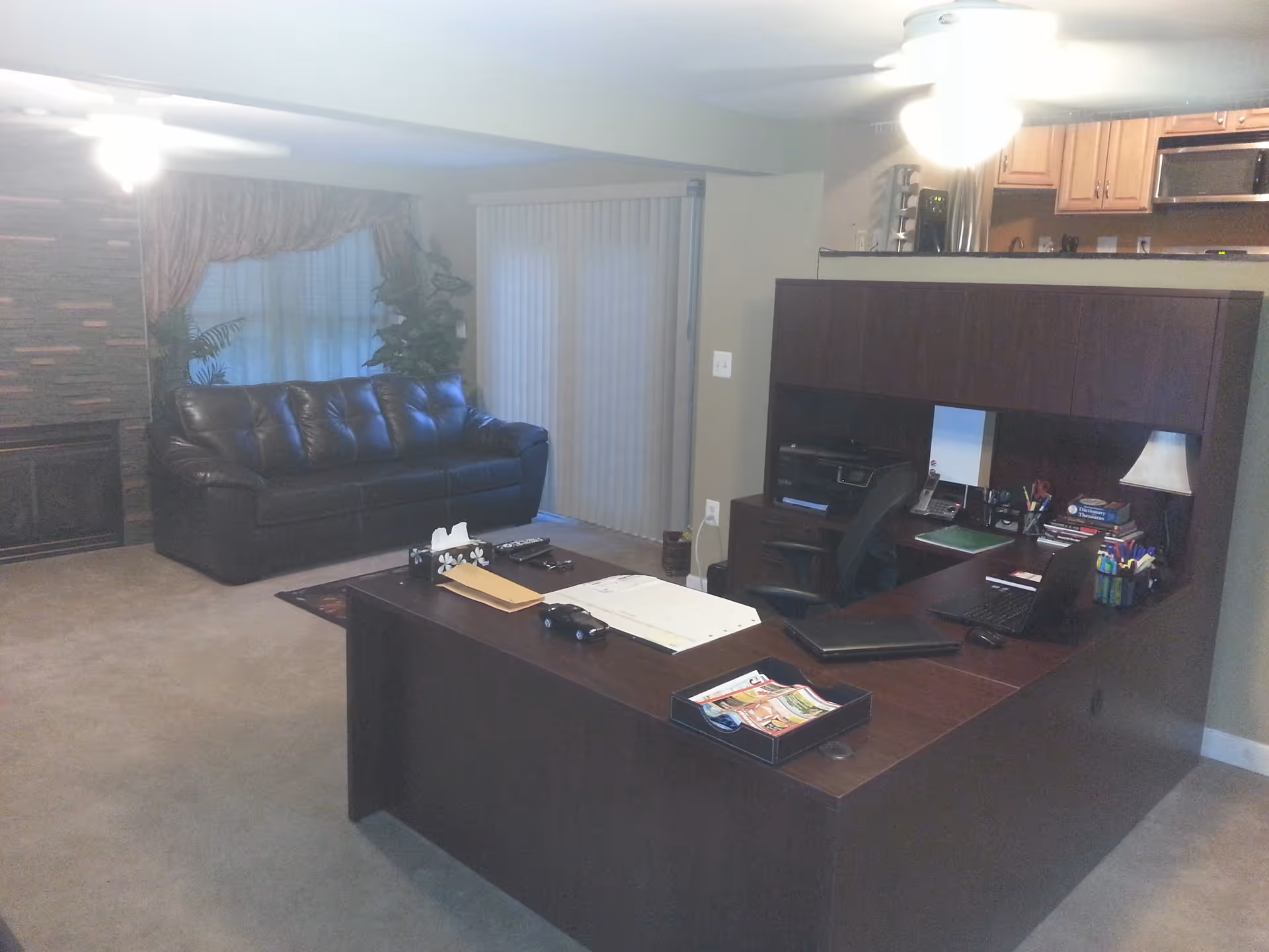 Open living room with a dark leather sofa and a large wooden desk/office workstation in the foreground, sliding glass doors and a kitchenette visible in the back.