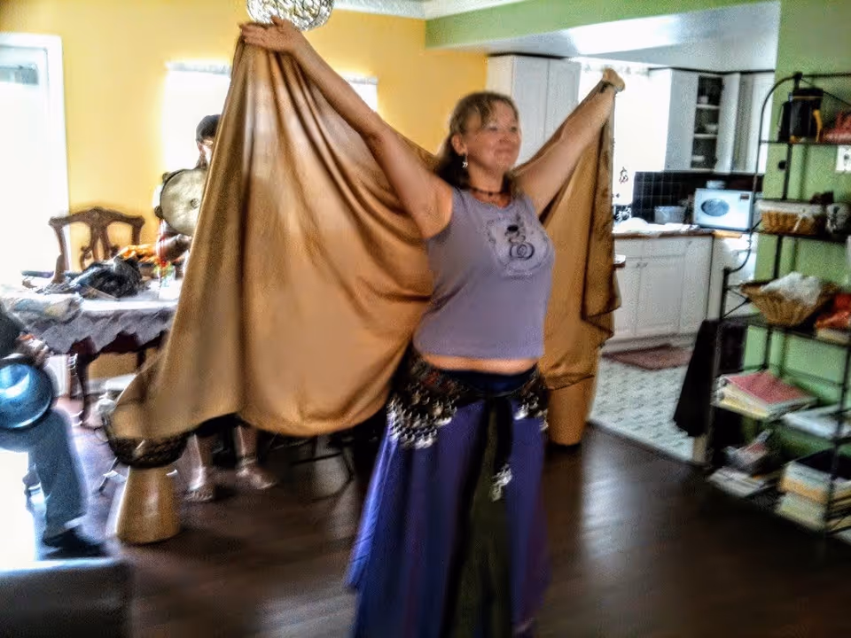 A woman indoors holding up a large golden fabric with both arms extended, wearing a purple top and a long skirt with decorative elements. In the background, there is a dining table with chairs and a kitchen area with white cabinets and black backsplash.