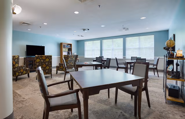 A well-lit common area with multiple wooden tables and chairs arranged throughout the room. There are large windows letting in natural light, a TV on a stand, a bookshelf, and a few patterned armchairs near the TV. A shelving unit with decorative items is visible on the right side of the room.