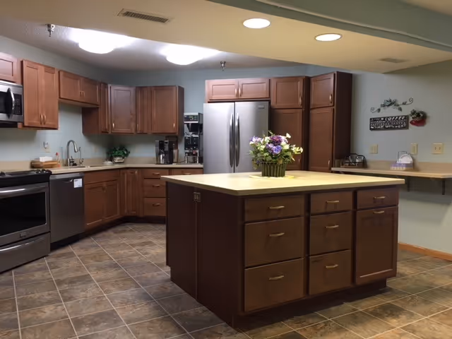 A spacious kitchen with wooden cabinets, a large central island with drawers, stainless steel appliances including a refrigerator, dishwasher, and oven. The floor is tiled, and there is a flower arrangement on the island. The walls are light-colored with a coffee-themed wall decoration and a small shelf with items on the right side.
