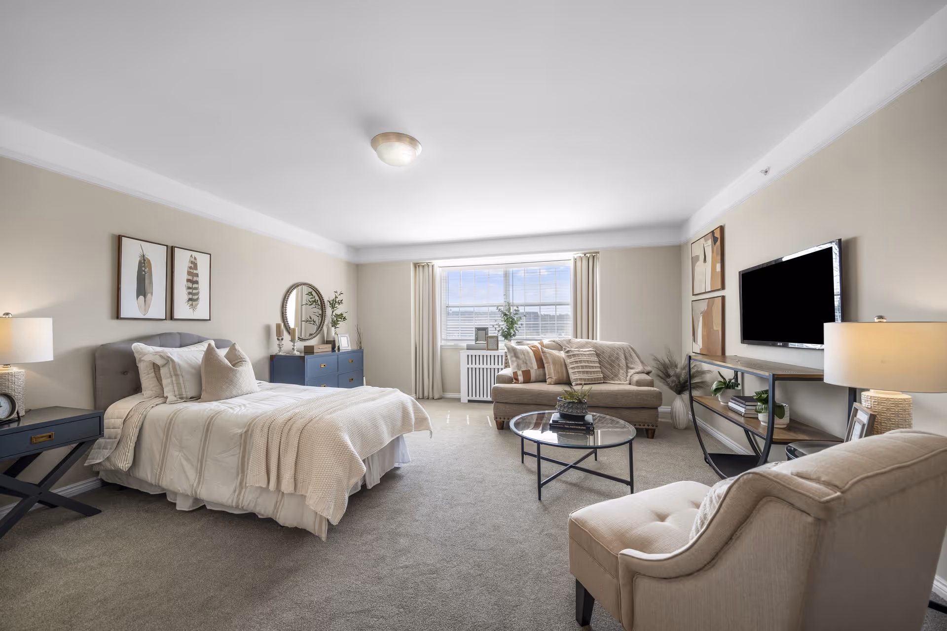 Spacious, well-lit studio-style bedroom and living area with a neatly made bed, upholstered seating, coffee table, and wall-mounted TV.