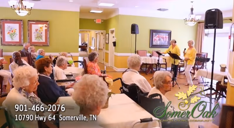 A group of elderly people seated in a common room, attentively watching two women standing and speaking or singing into microphones. The room has green walls, framed artwork, and tables covered with white tablecloths.