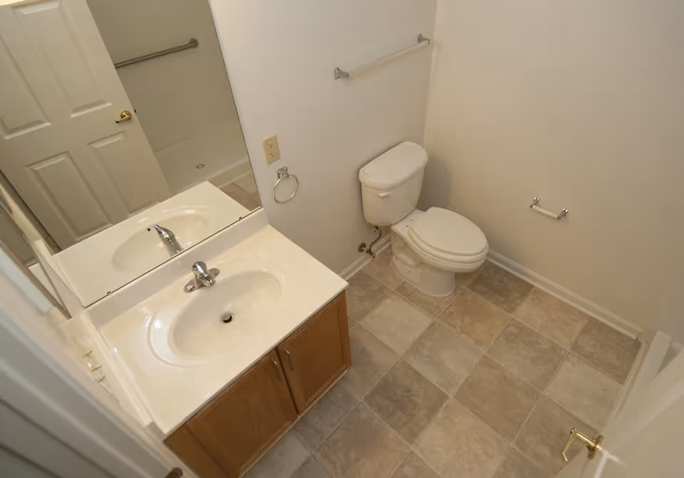 A small bathroom with a white toilet, a sink with a countertop and wooden cabinet below, a large mirror above the sink, beige and gray tiled floor, and white walls with a towel rack and toilet paper holder.