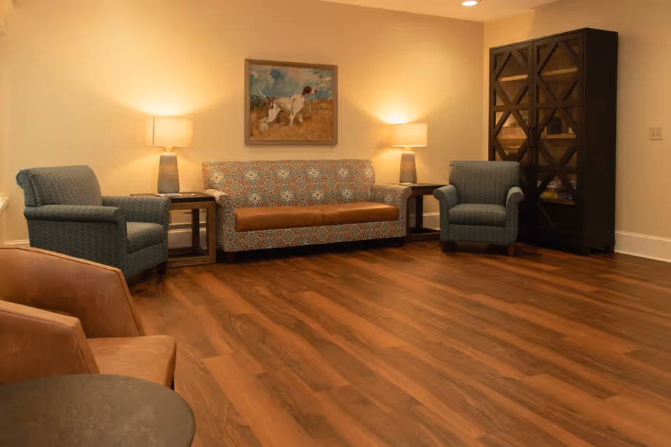 A cozy living room with wooden flooring, featuring a patterned sofa with a brown leather seat cushion, two blue armchairs, two side tables each with a lamp, a dark wooden cabinet with glass doors, and a painting of a dog on the wall.