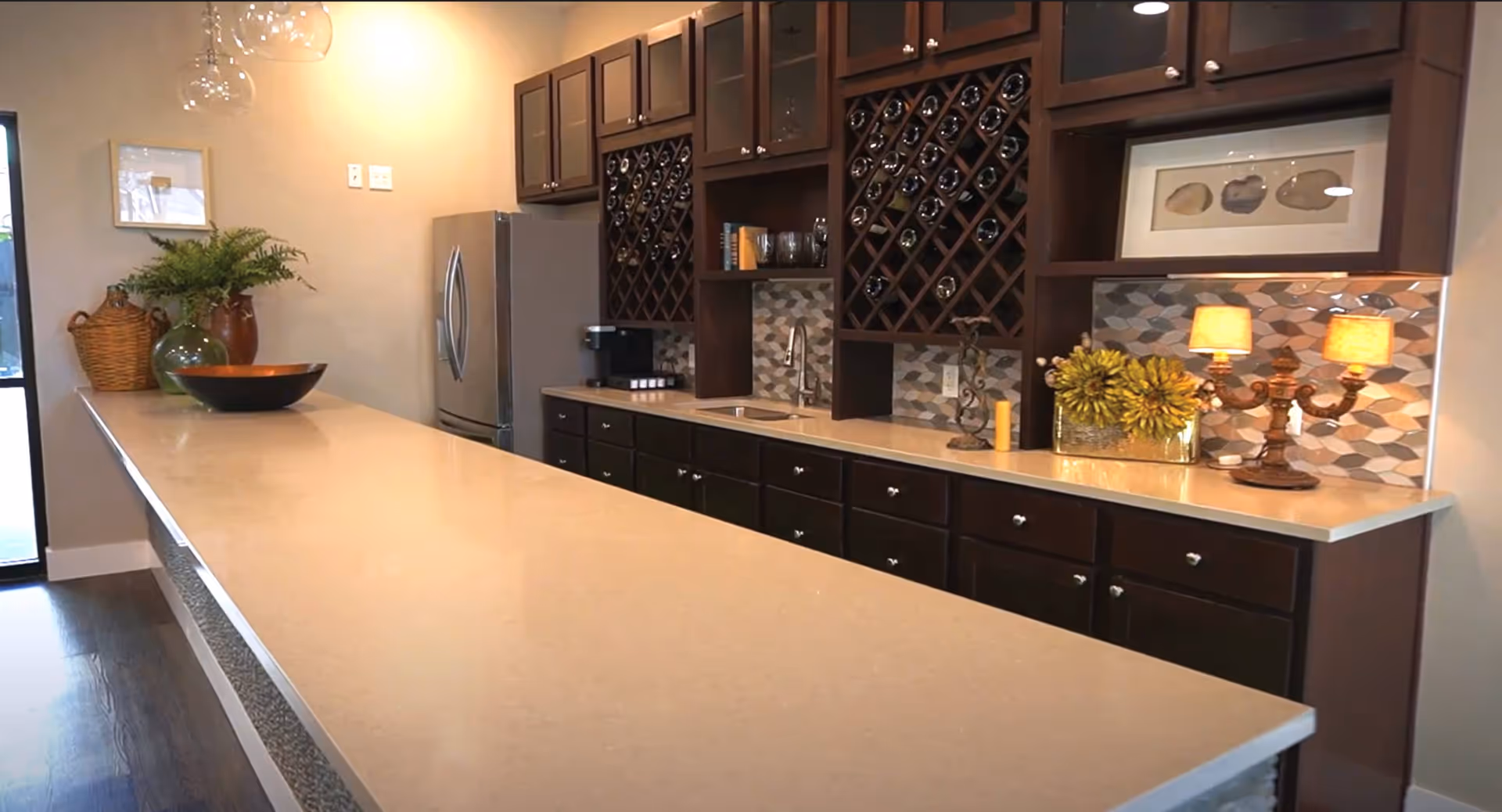 Modern kitchen area with a long beige countertop island, dark wooden cabinets, a stainless steel refrigerator, wine racks, decorative plants, a table lamp, and framed artwork on the wall.