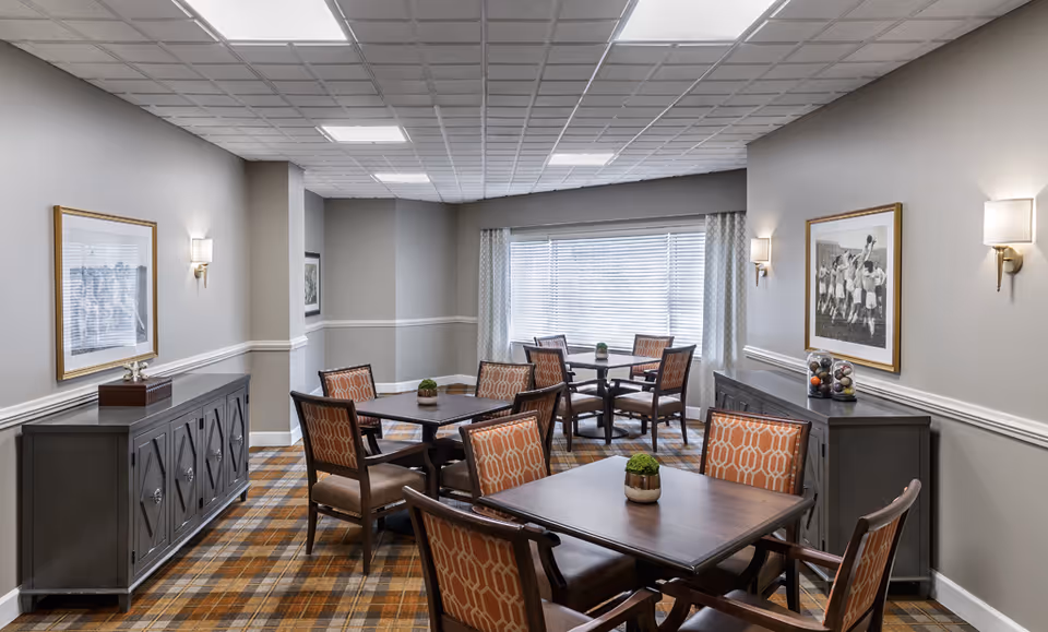Communal dining room with several square wooden tables surrounded by patterned upholstered chairs, sideboards, framed artwork, and a large window.