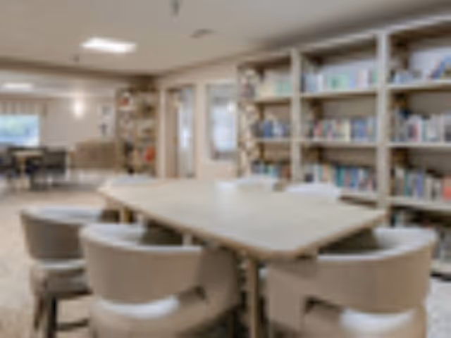 A well-lit interior room featuring a large rectangular table surrounded by six cushioned chairs. In the background, there are bookshelves filled with books and a seating area with additional chairs near windows.