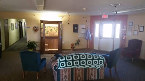 A cozy seating area in a healthcare facility with a patterned couch draped with a green and white crocheted blanket, two blue armchairs, and a red armchair. The room has beige walls, a carpeted floor, a hallway leading to other rooms, a door with a window, and some plants and wall decorations.