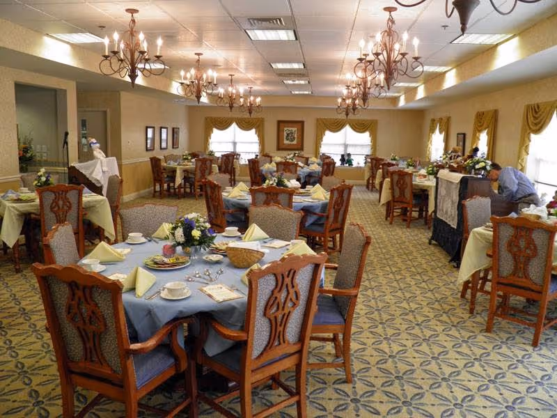 Spacious dining room with round tables set with place settings, floral centerpieces, and chandeliers overhead.