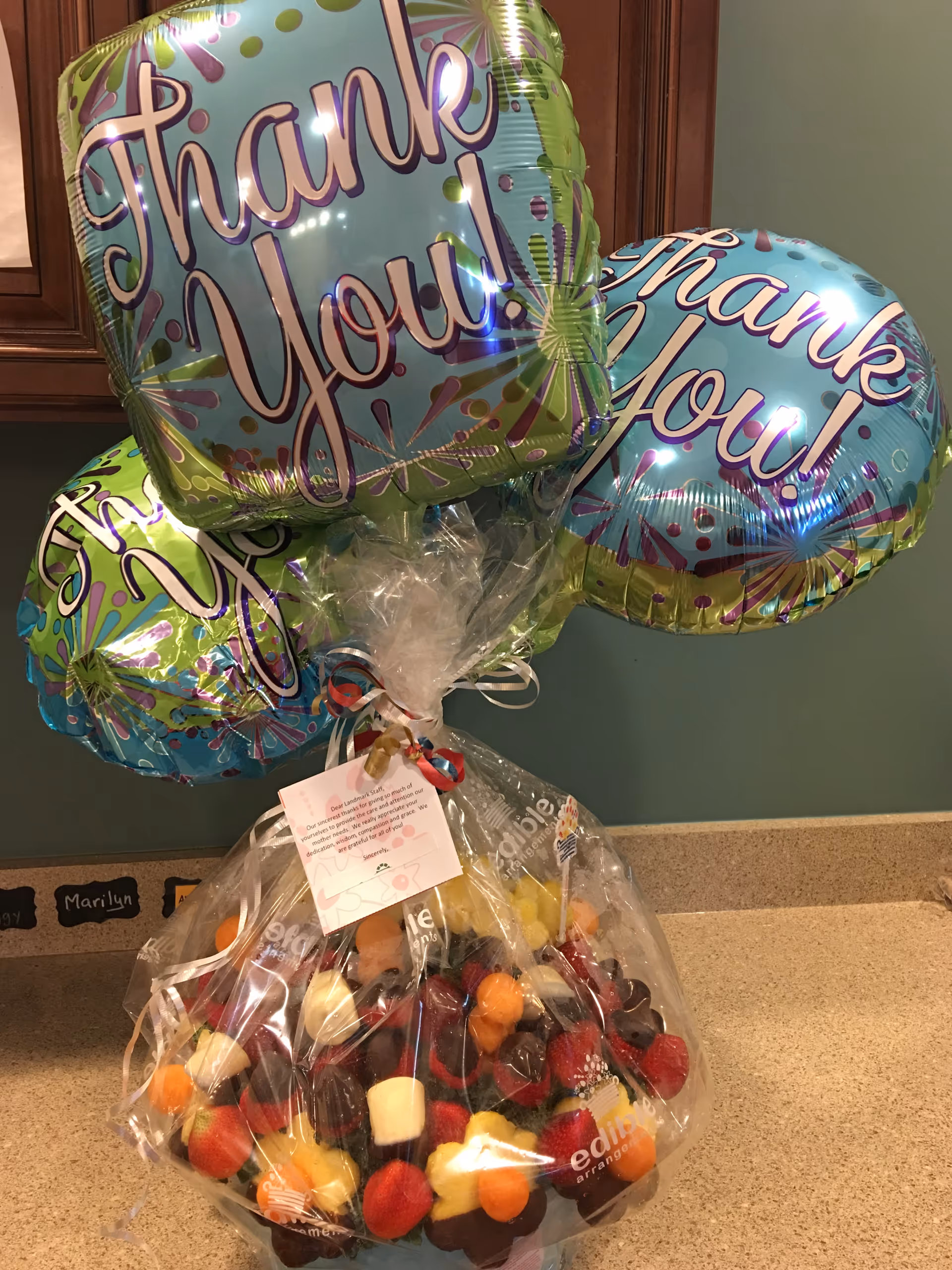A wrapped fruit bouquet on a kitchen counter topped with three "Thank You!" balloons.