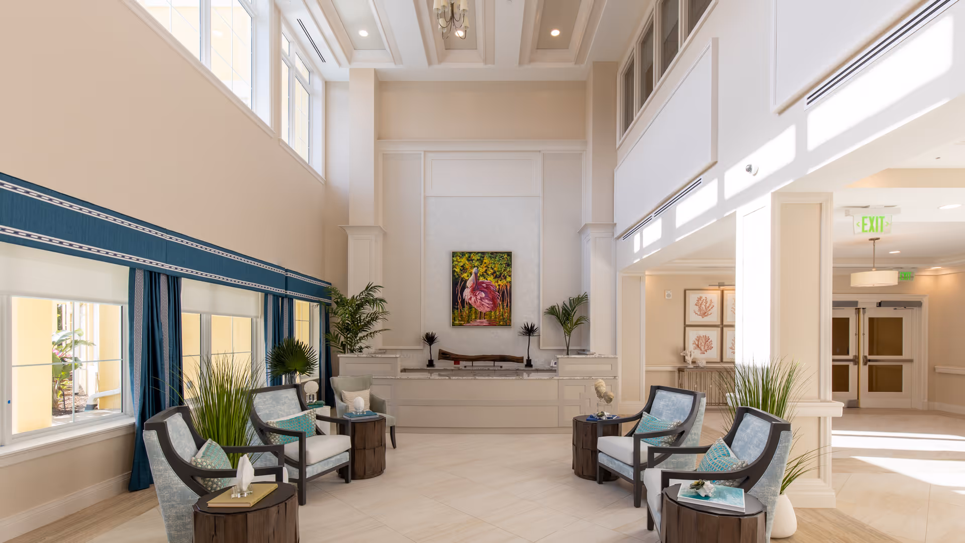 A bright and spacious living room area with high ceilings and large windows letting in natural light. The room features six cushioned armchairs arranged in pairs around small wooden side tables, with decorative plants placed throughout. A colorful painting of a flamingo hangs on the far wall above a low mantel, and the walls and floor are in neutral tones. The space has a welcoming and elegant atmosphere.