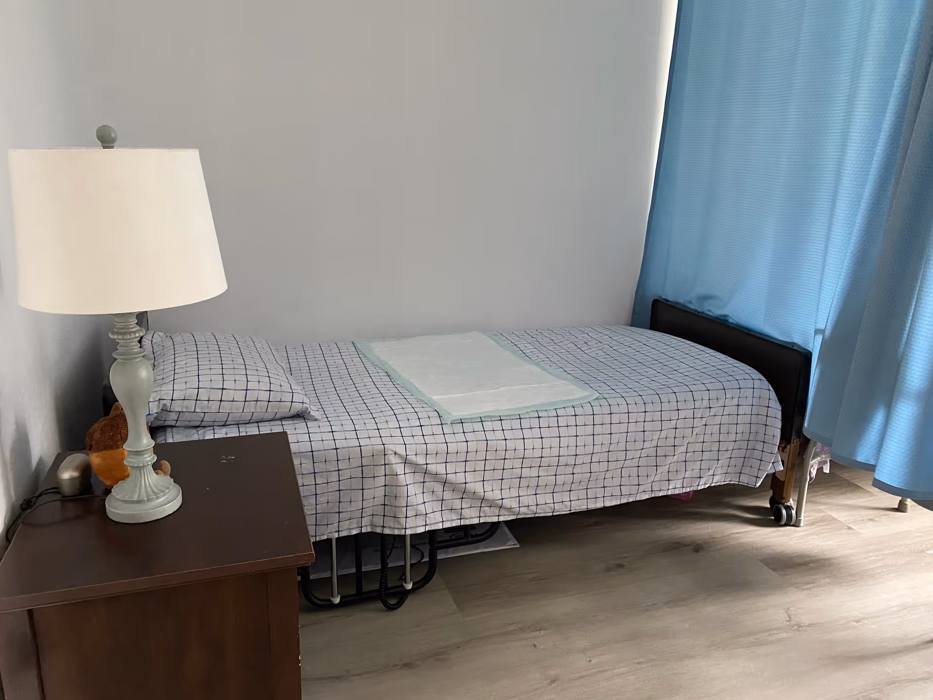 A simple bedroom with a single bed covered in a checkered bedspread and a white absorbent pad on top. Next to the bed is a wooden nightstand with a white lamp and a small stuffed animal. Blue curtains cover the window on the right side of the room, and the floor is light-colored wood.