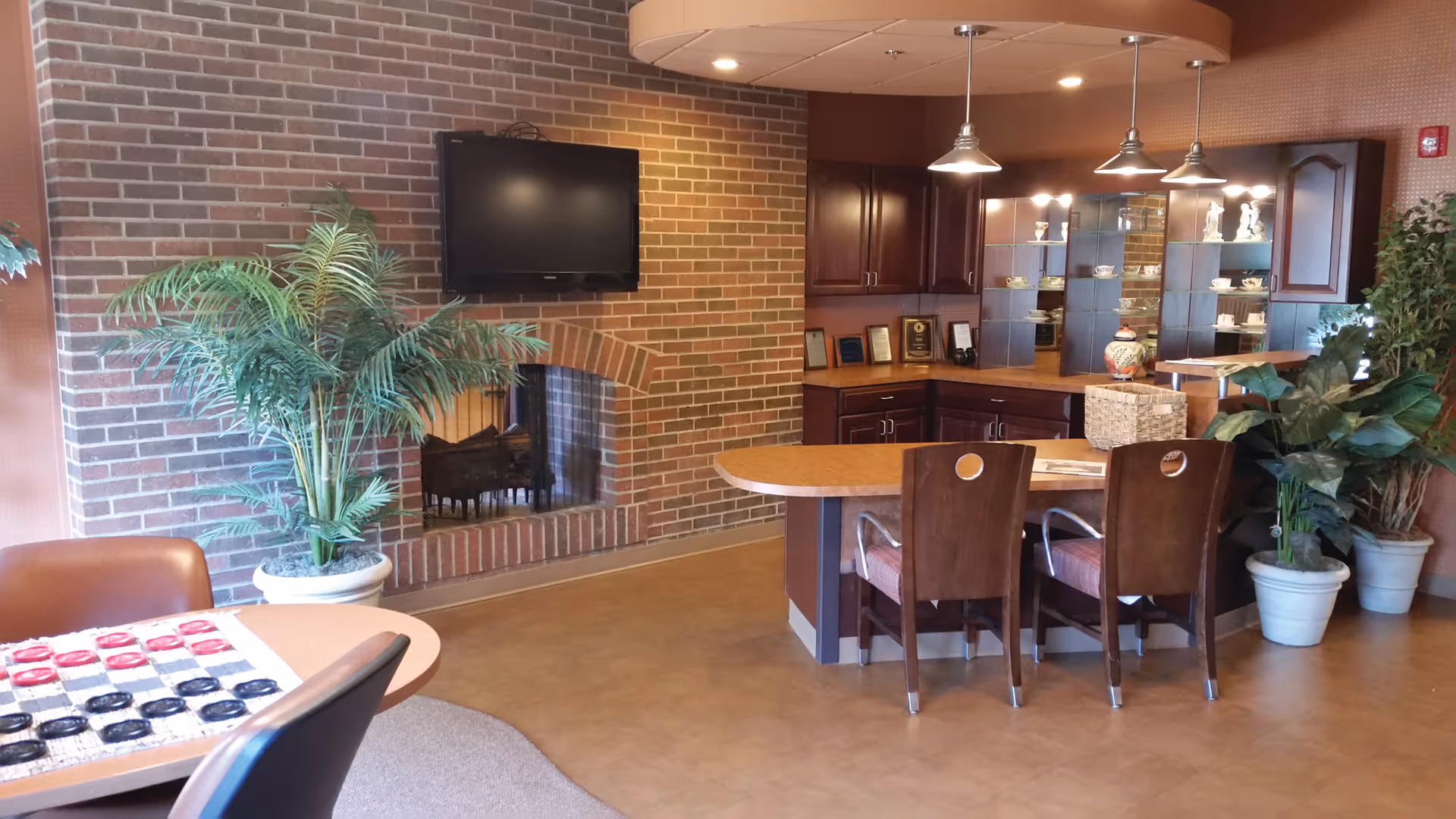 Interior view of a common area featuring a brick fireplace with a mounted flat screen TV above it, a round table with a checkers game, a counter with three wooden chairs, and several potted plants. The background includes wooden cabinetry with glass shelves displaying decorative items and plaques.