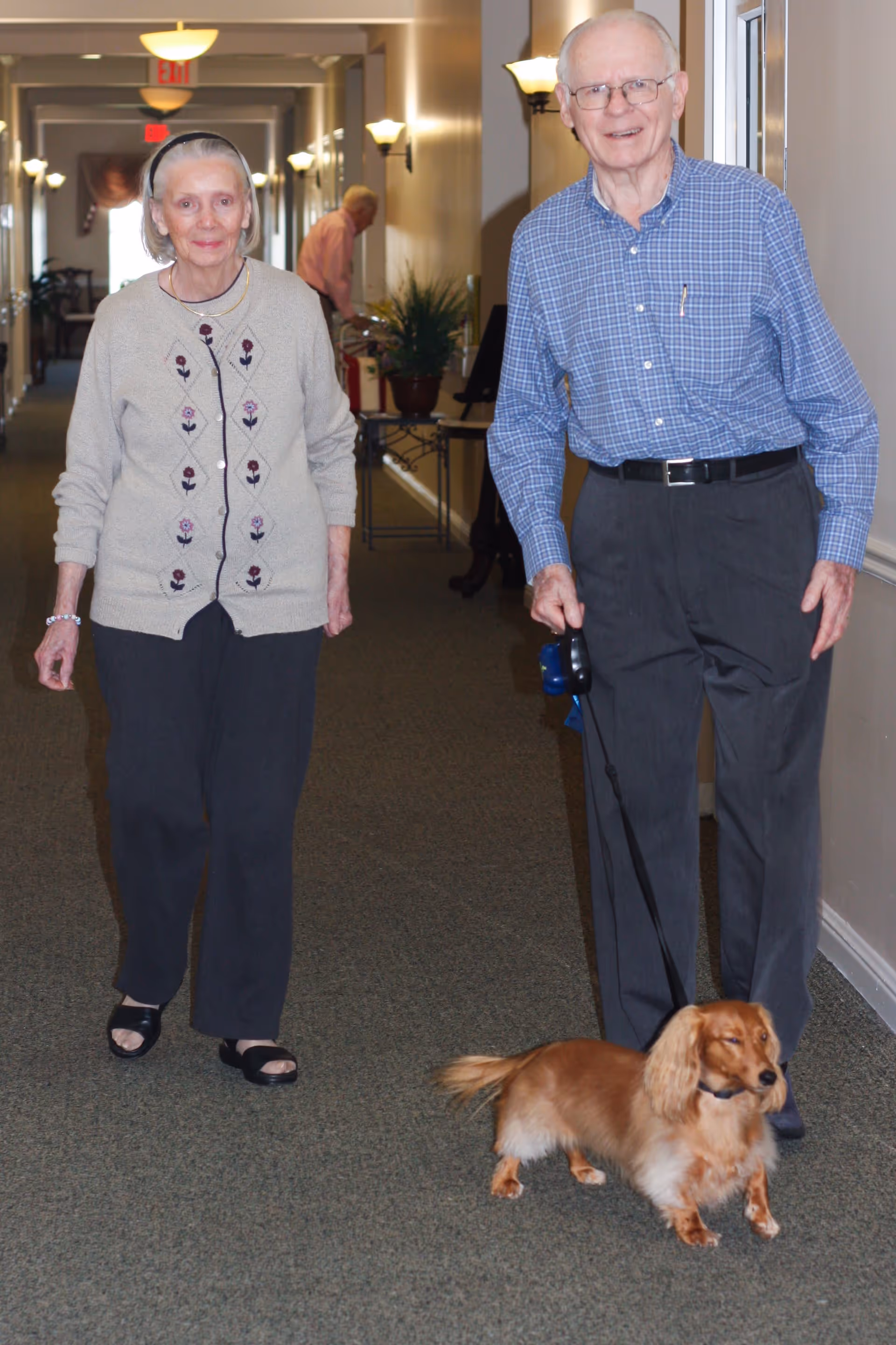 An elderly man and woman walking down a carpeted hallway inside a senior living facility. The man is holding a leash attached to a small brown dog. The hallway is well-lit with wall sconces and ceiling lights, and there is a person in the background near a table with plants.