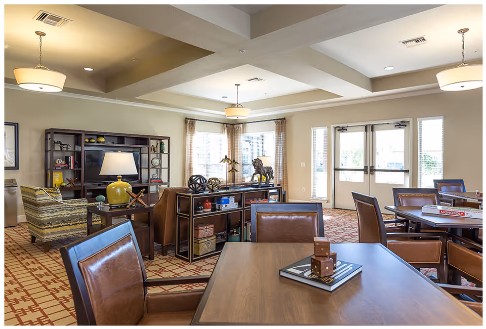 A spacious and well-lit common area in a senior living facility featuring multiple tables with brown leather chairs, a bookshelf with decorative items, a TV, and large windows with curtains allowing natural light to fill the room. The carpet has a red and beige geometric pattern, and there are ceiling lights providing additional illumination.