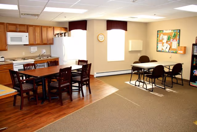 A well-lit room featuring a kitchen area with wooden cabinets, a white refrigerator, microwave, stove, and sink. In front of the kitchen is a wooden dining table with six chairs. Adjacent to this area is a small table with four chairs, a bulletin board on the wall, and a bookshelf filled with books and games. Two windows with maroon valances allow natural light into the room.