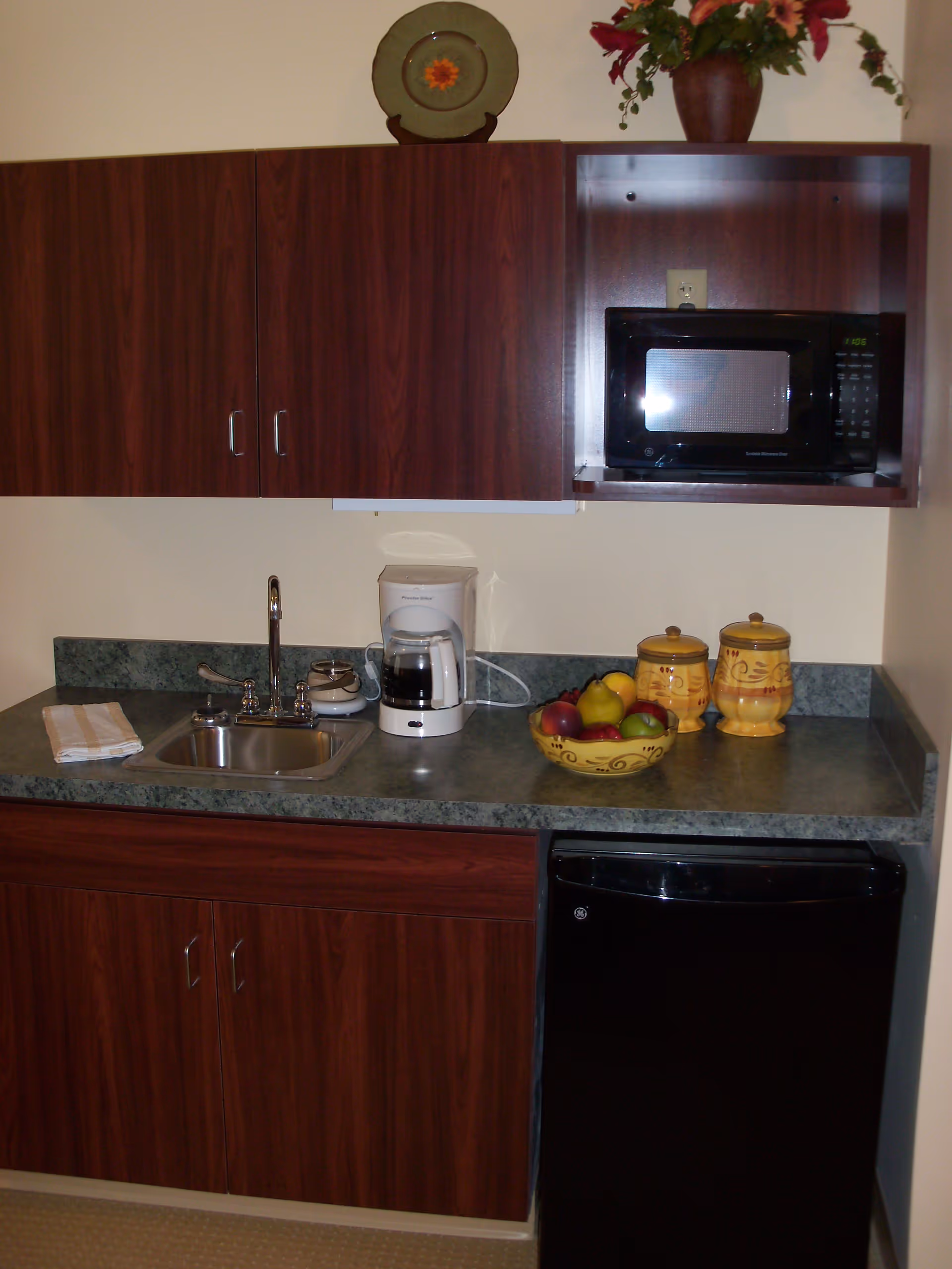 A small kitchen area with dark wood cabinets, a green countertop, a stainless steel sink with a faucet, a white coffee maker, a bowl of assorted fruit, two yellow decorative jars, a black microwave, and a black mini refrigerator. There is a decorative plate and a flower vase on top of the upper cabinets.