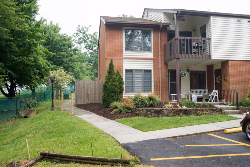 Exterior view of a two-story residential building with brick and siding. There is a small balcony on the upper floor and a porch area with seating on the ground floor. The building is surrounded by a well-maintained lawn, shrubs, and trees. A paved walkway leads to the entrance, and a parking area is visible in the foreground.