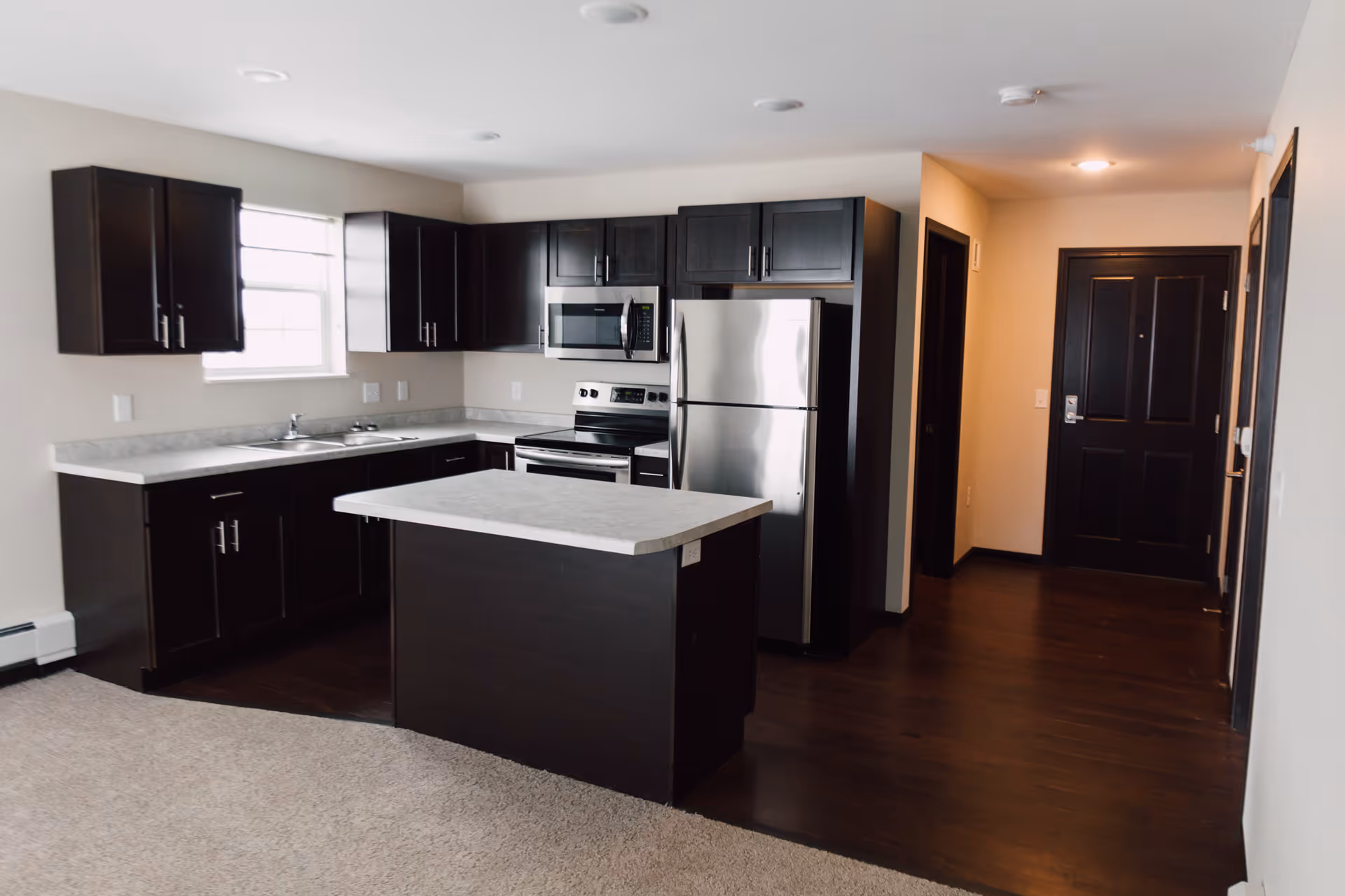 Modern kitchen area with dark wood cabinets, a stainless steel refrigerator, microwave, and stove. There is a kitchen island with a light-colored countertop, a window above the sink, and a dark wood floor leading to a black front door in the background.