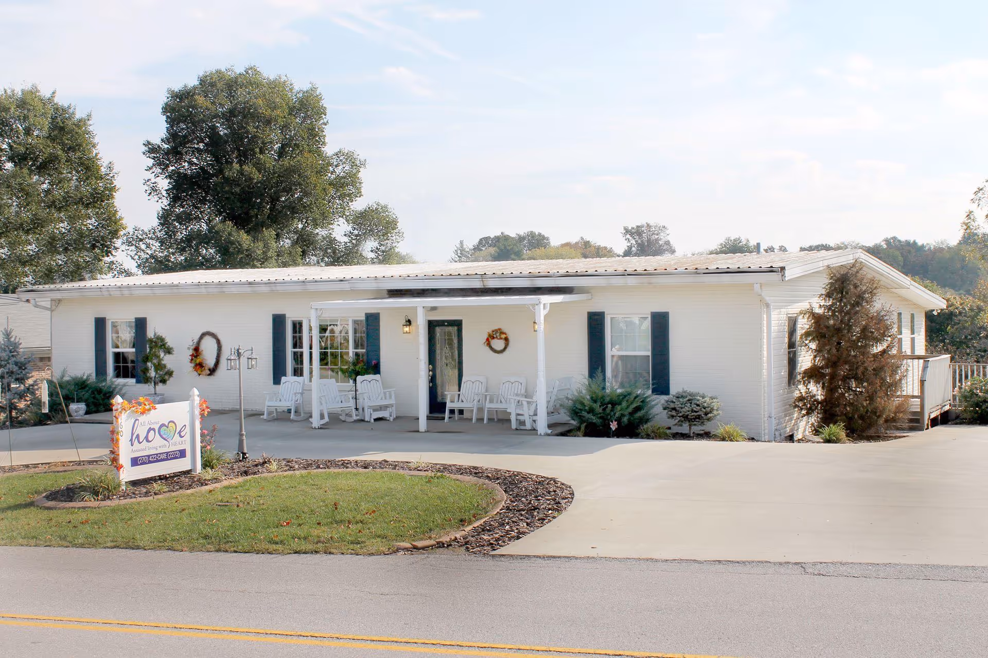 Front view of a single-story white assisted living building with a covered porch, chairs, landscaping, and a sign by the driveway.