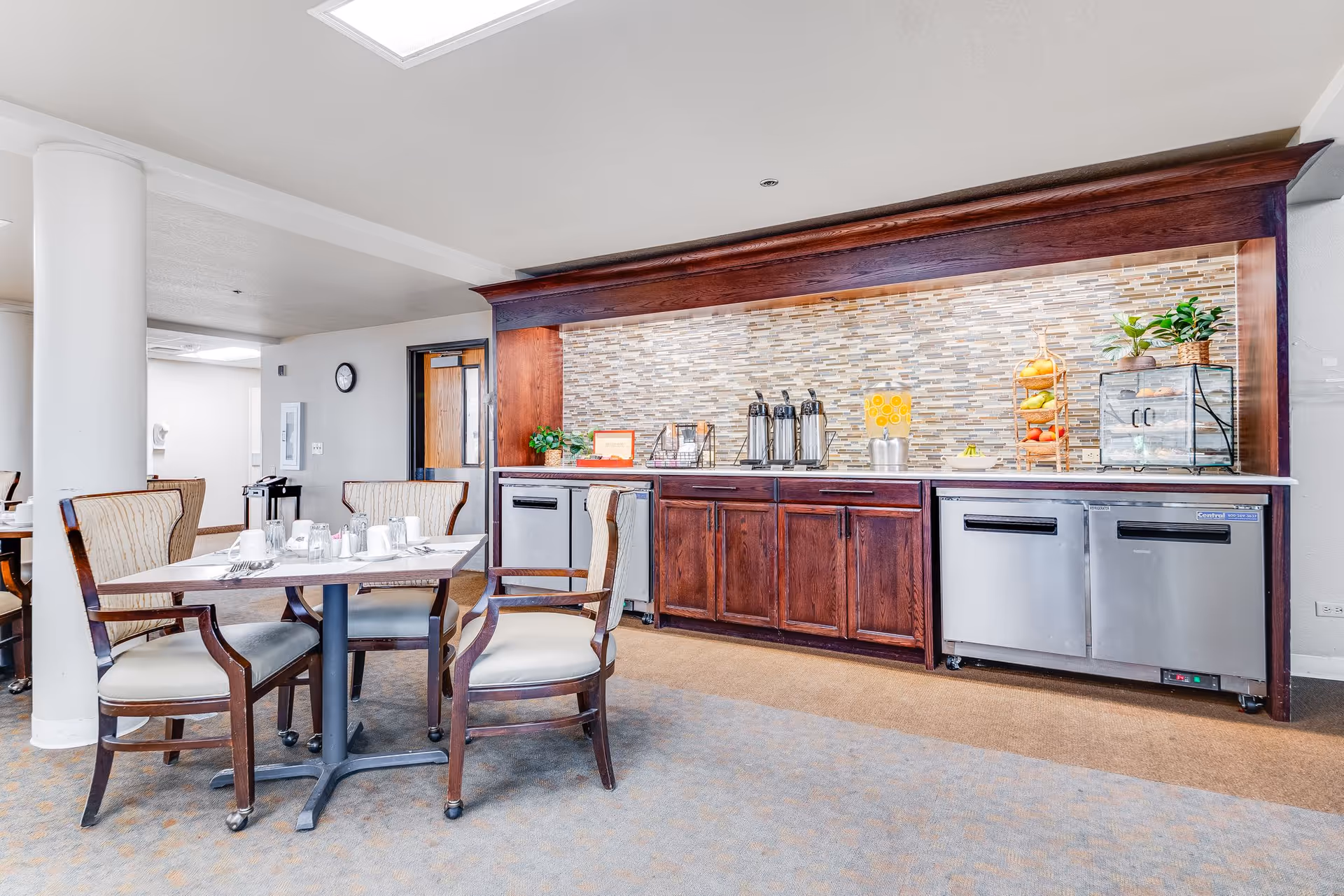 Dining area with a table and chairs in front of a wooden service counter stocked with beverage dispensers and a fruit display.