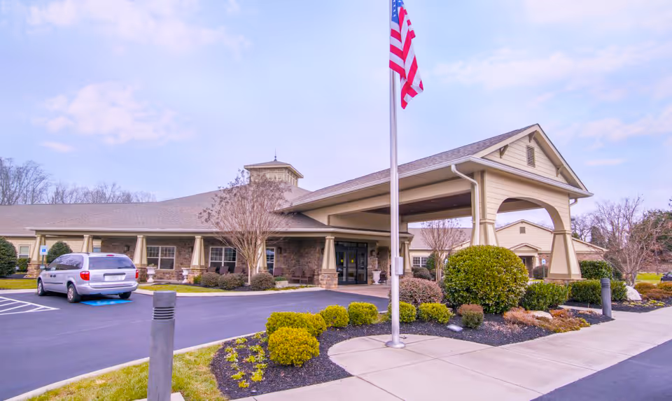 Exterior view of Morning Pointe of Lenoir City senior living facility showing a single-story building with a covered entrance, landscaped bushes, an American flag on a flagpole, and a parked silver minivan in a handicapped parking space.