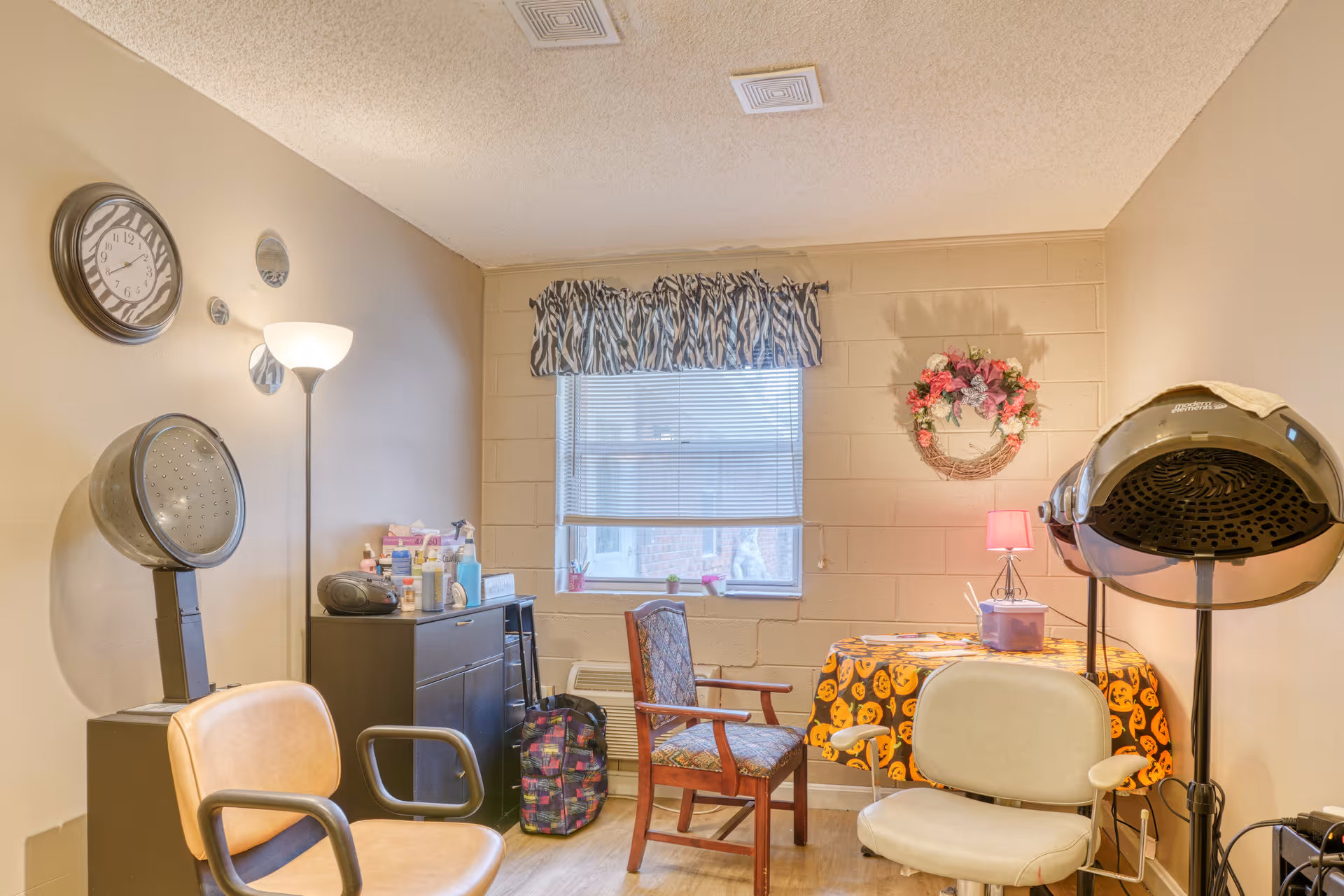 A small room with beige walls and a window with zebra-patterned valance. The room contains two salon hair dryers, a floor lamp, a clock on the wall, a small black cabinet with various bottles and items on top, a wooden chair, and a table covered with a Halloween-themed tablecloth with a small pink lamp and a container on it.