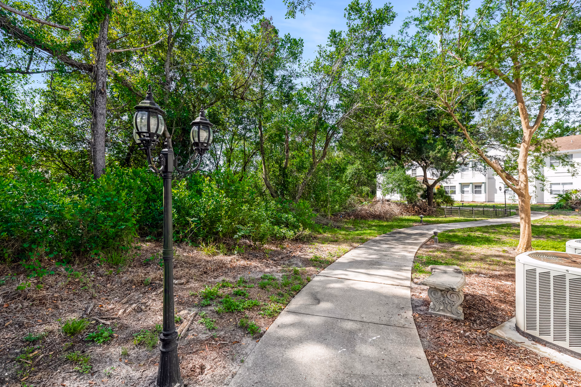A winding concrete pathway through a green outdoor area with trees and bushes on either side. There is a vintage-style black street lamp on the left and a stone bench on the right near an air conditioning unit. In the background, a white building is partially visible under a clear blue sky.