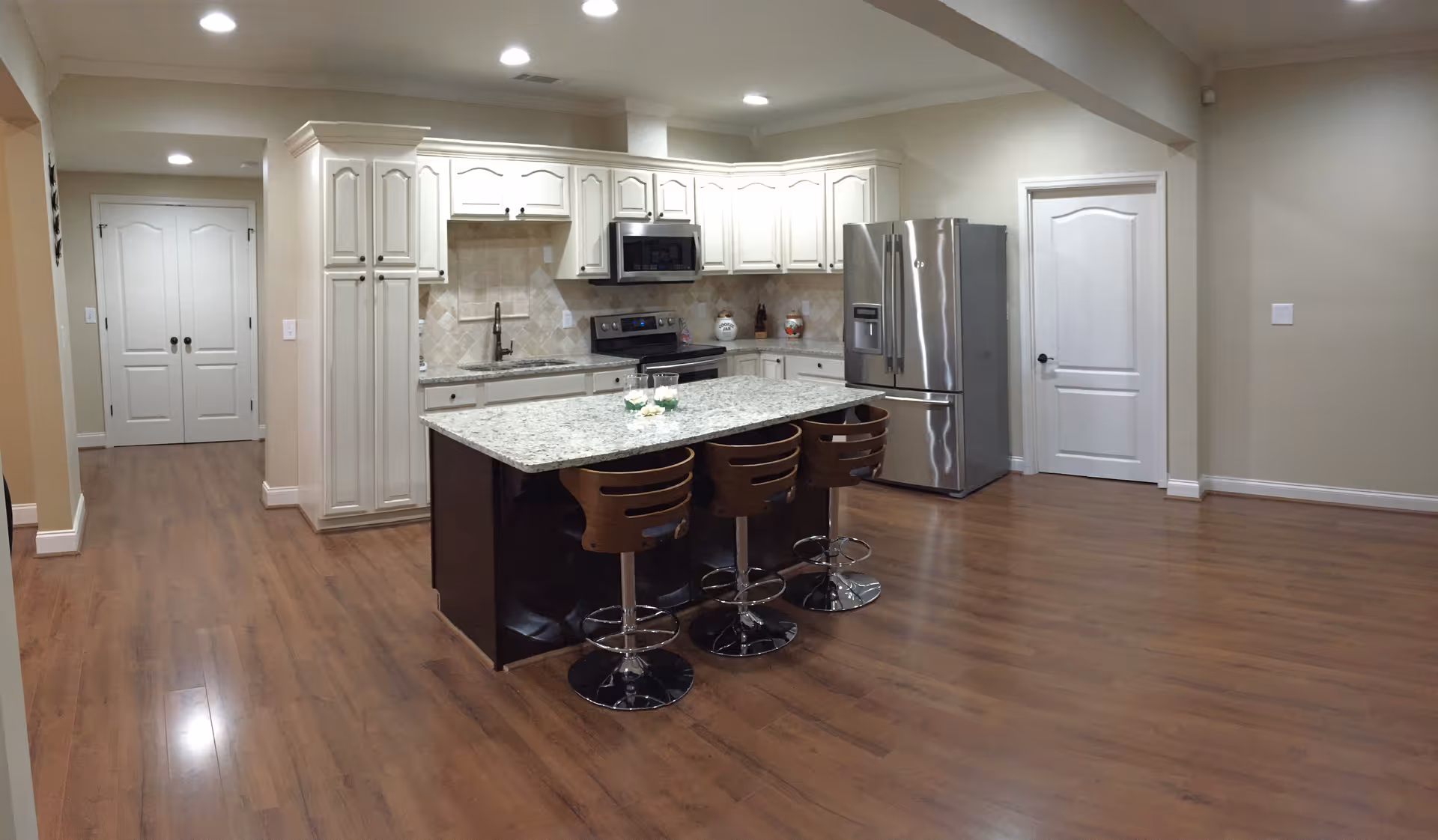 Open modern kitchen with a granite island and three wooden bar stools, white cabinetry, stainless steel appliances, and wood flooring.