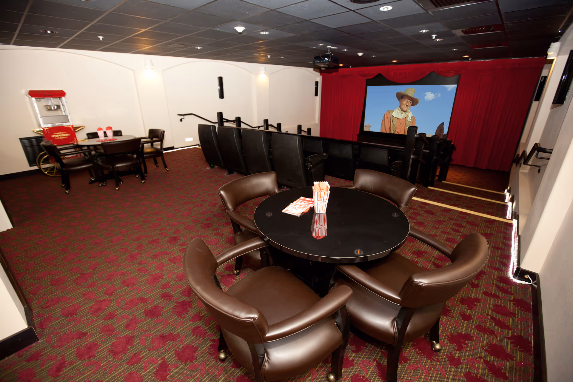 A community screening room with round tables and leather chairs, a popcorn machine, and a movie screen showing a cowboy.