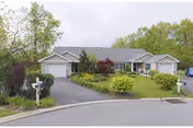 Single-story residential building with two attached garages, surrounded by green trees and shrubs, with a driveway and mailboxes in front.
