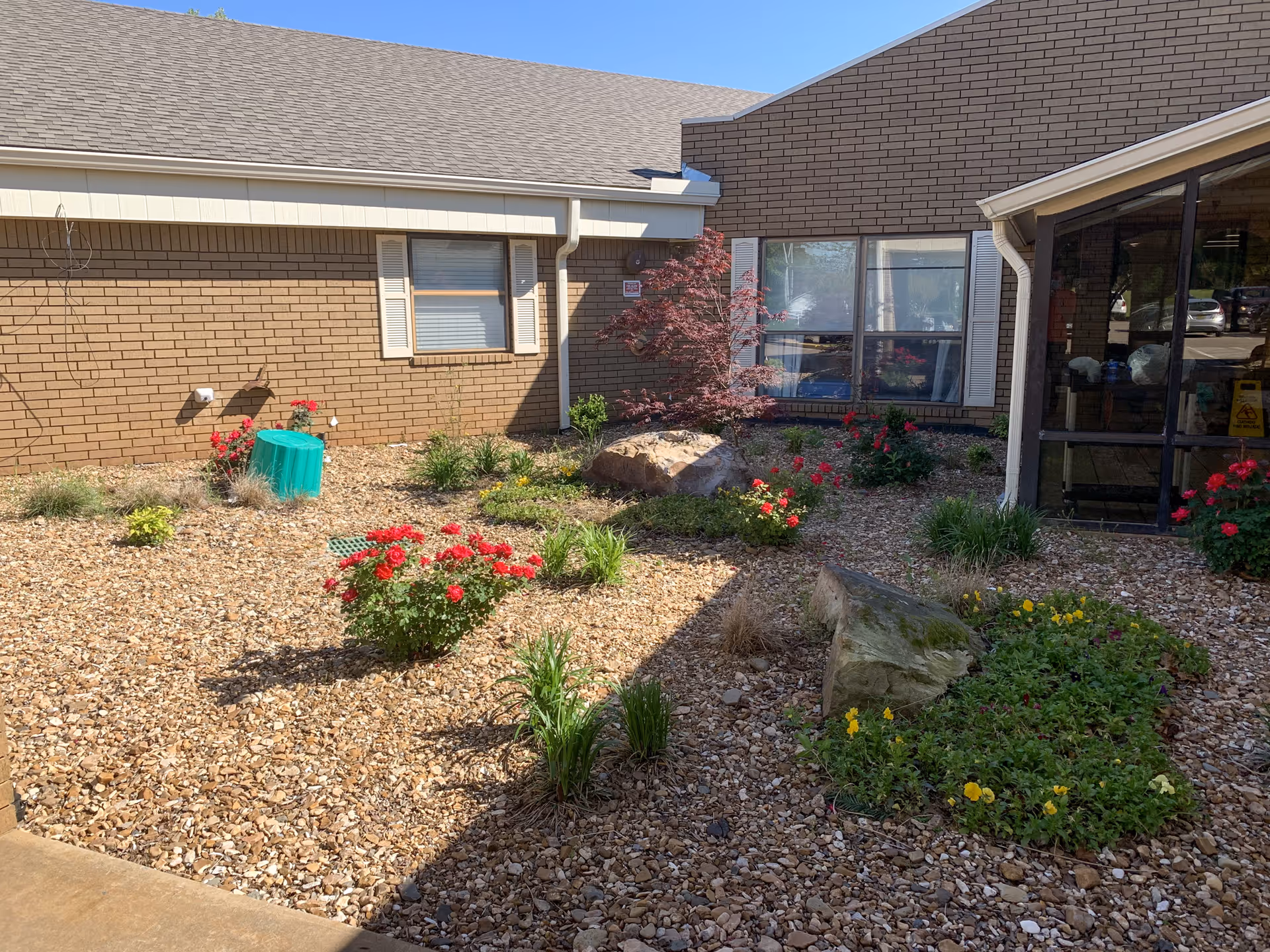 Outdoor garden area at Oaks Healthcare Center featuring a variety of plants and flowers, including red and yellow blooms, surrounded by small rocks and mulch. The garden is adjacent to a brick building with windows and a glass-enclosed entrance.