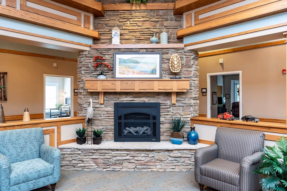 A cozy common area with a stone fireplace surrounded by wooden trim. The fireplace mantel holds decorative items including a framed landscape painting, a lantern, and vases. Two upholstered armchairs, one blue patterned and one gray patterned, are positioned on either side of the fireplace. There are small plants and decorative objects on the hearth and nearby surfaces. The walls are beige with wooden accents, and doorways lead to other rooms in the background.
