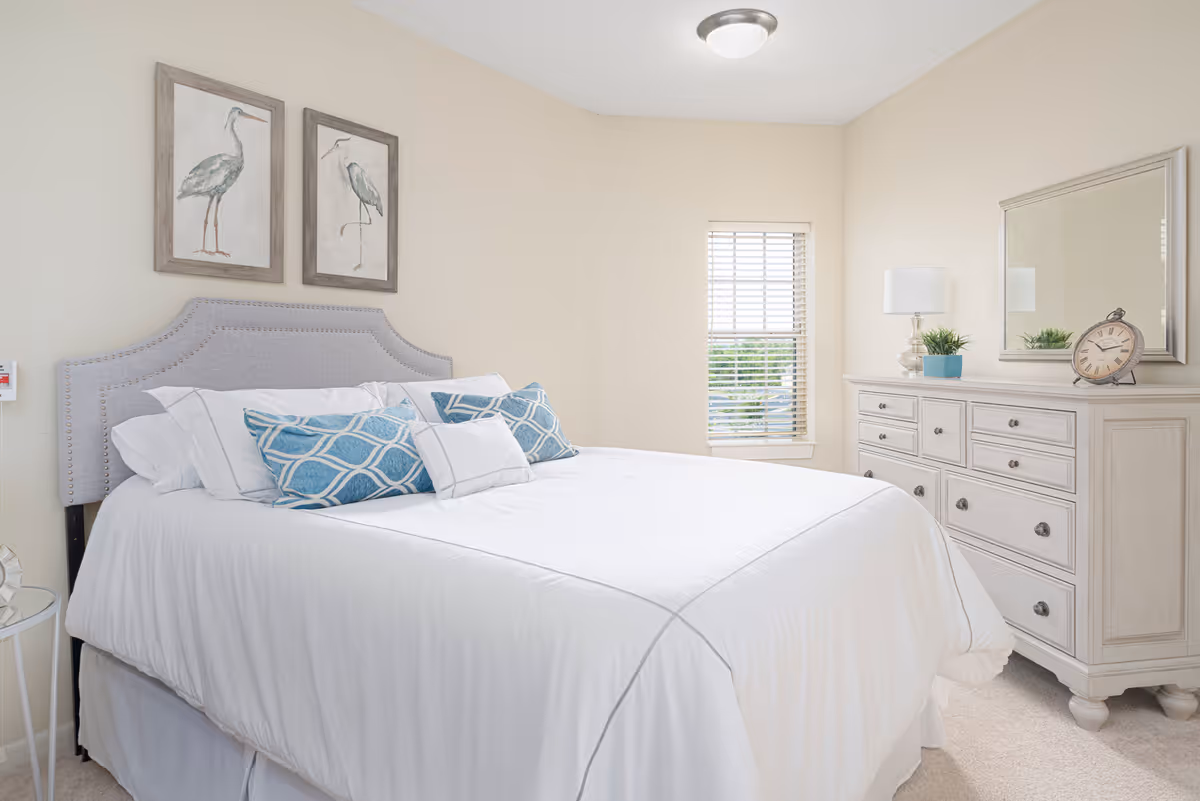A bright bedroom with a large bed featuring a gray upholstered headboard, white bedding, and decorative blue and white pillows. Above the bed are two framed pictures of birds. To the right, there is a white dresser with multiple drawers, a table lamp, a small plant, a decorative clock, and a large mirror hanging above it. A window with white blinds is on the wall behind the dresser.