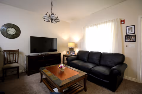 A cozy living room with a black leather sofa, a wooden coffee table with a small flower arrangement, a wooden TV stand with a flat-screen television, a wooden chair, a wall clock, a table lamp on a side table, and framed photos on the wall near a window with white curtains.