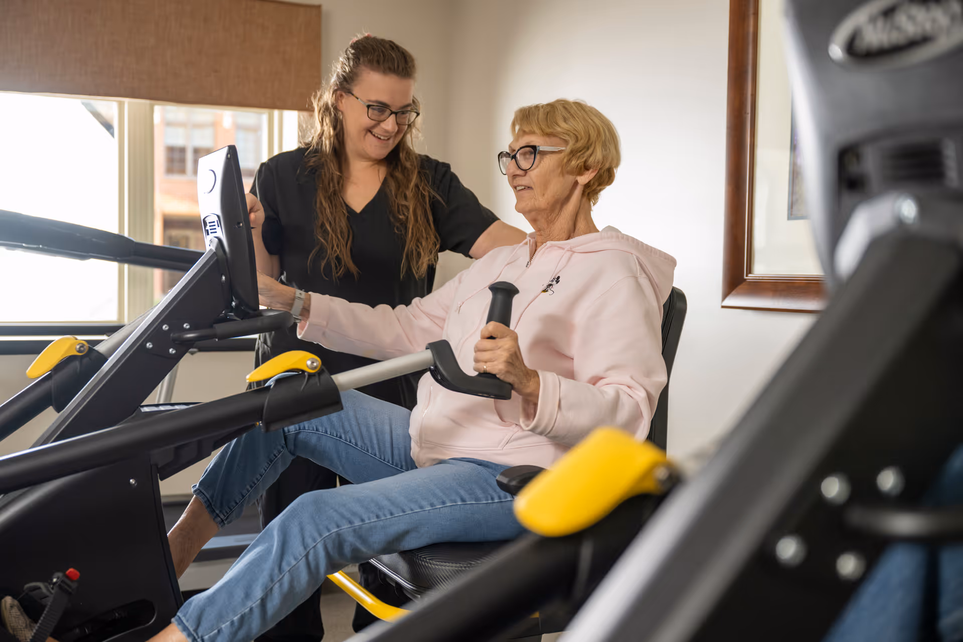 An elderly woman wearing glasses and a pink hoodie is exercising on a seated fitness machine while a younger woman in black scrubs stands beside her, smiling and offering support in a well-lit room with a window and framed artwork on the wall.