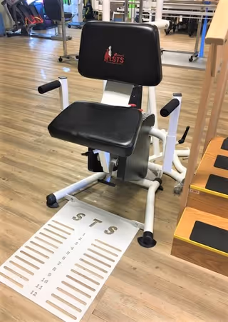 A black and white exercise machine with a padded seat and backrest, positioned on a wooden floor. The backrest has the logo 'STS' on it. Next to the machine is a wooden step platform with black non-slip pads. The background shows more exercise equipment in a fitness or rehabilitation room.