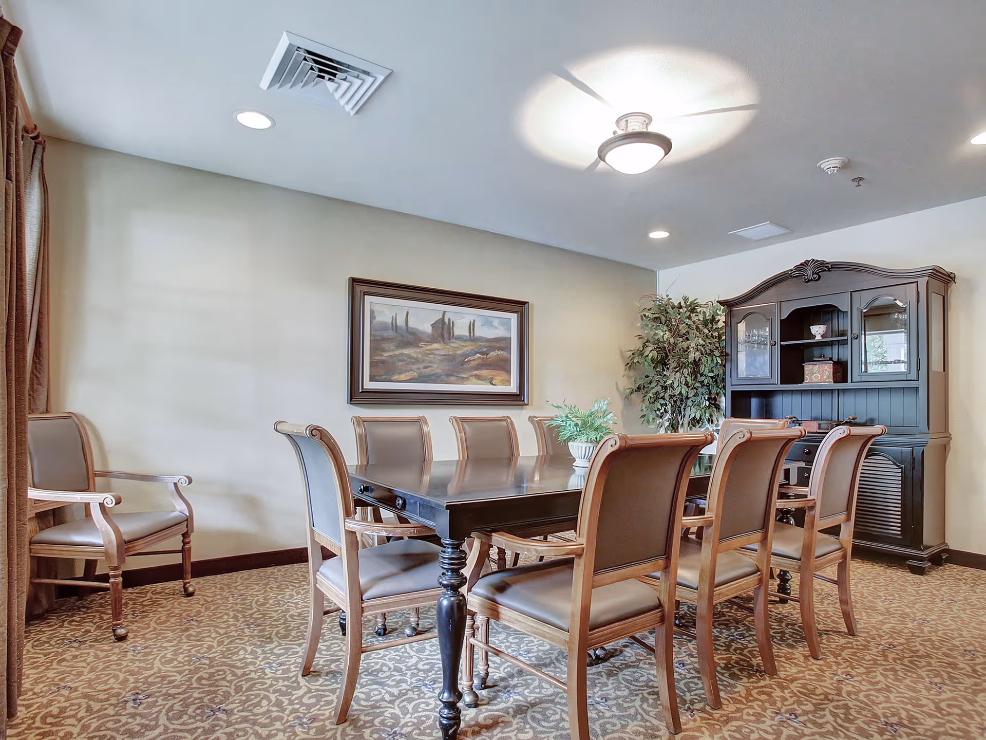 A dining room with a large rectangular wooden table surrounded by eight wooden chairs with cushioned seats and backs. There is a framed landscape painting on the wall, a large dark wooden cabinet with glass doors and shelves, and a potted plant in the corner. The room has patterned carpet flooring and a ceiling light fixture.