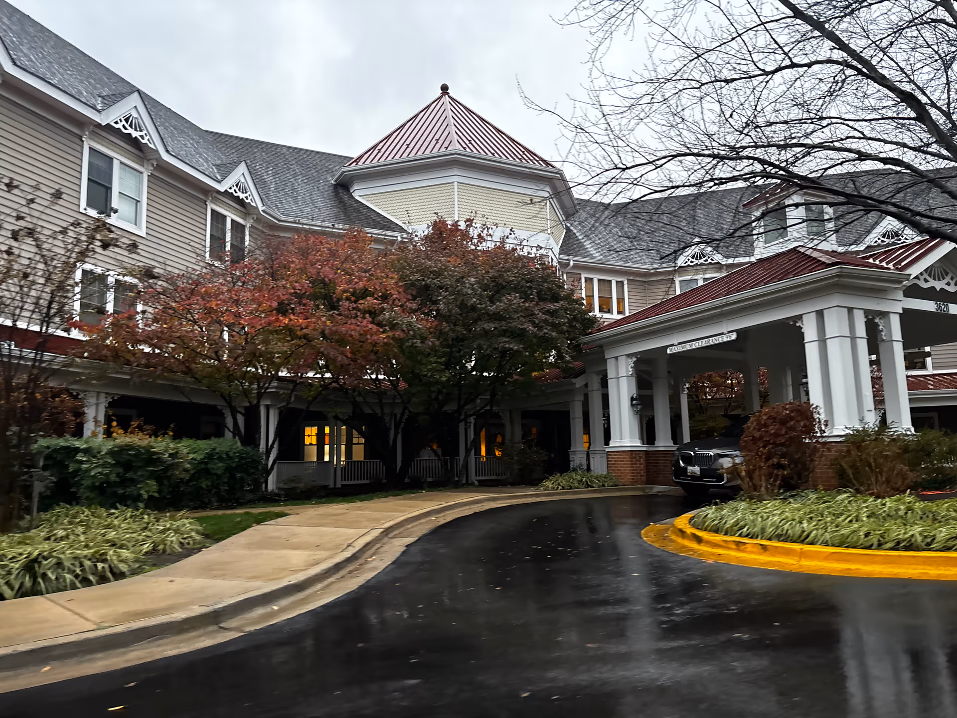Front entrance of a senior living building with a covered porte-cochère, autumn trees, and a wet driveway.