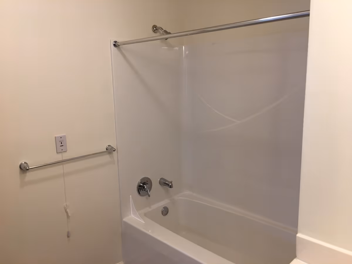 A clean bathroom featuring a white bathtub with a showerhead and a chrome faucet. There is a chrome towel bar mounted on the wall next to the bathtub, and a pull cord hanging from a switch above the towel bar. The walls are painted a light beige color.